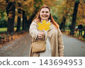 Portrait of pretty young 30s woman in evening autumn park, smiling. Autumn landscape, calmness, tranquility concept. Pretty female holding leaf on nature background. High quality photo. 143694953