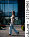 Pretty Woman Walking Near Glass Office Building Wearing Beige Blazer And Jeans. Casual Business Lifestyle Concept Female Urban Fashion Style 143694966