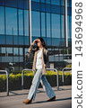 Beautiful Woman Walking In City Wearing Beige Blazer And Jeans Near Modern Office Building. Casual Business Lifestyle Concept Female Urban Fashion 143694968