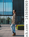 Pretty Woman Walking Near Glass Office Building Wearing Beige Blazer And Jeans. Casual Business Lifestyle Concept Female Urban Style 143694969