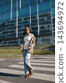 Beautiful Woman Walking Across Crosswalk Near Modern Office Building Wearing Beige Blazer And Jeans. Casual Business Lifestyle Concept Female Urban 143694972