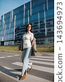 Attractive Woman Walking On Crosswalk Near Modern Office Building Wearing Beige Blazer And Jeans. Casual Business Lifestyle Concept Female Urban Style 143694973