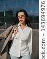Pretty Woman In White Shirt And Glasses Holding Jacket Near Modern Office Building. Casual Business Lifestyle Concept Female Urban Portrait 143694976