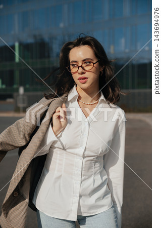 Pretty Woman In White Shirt And Glasses Holding Jacket Near Modern Office Building. Casual Business Lifestyle Concept Female Urban Portrait Pretty Woman In White Shirt And Glasses Holding Jacket Near Modern Office Building. Casual Business Lifestyle Concept Female Urban Portrait 143694976