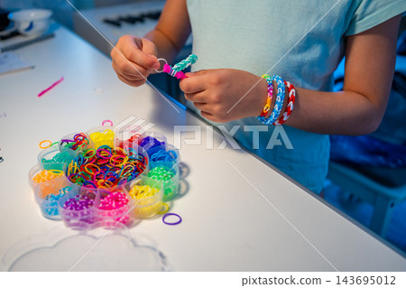 Child makes a bracelet from colorful rubber bands using a mini loom at home. Creative activity helps develop fine motor skills, hand eye coordination and concentration through play 143695012