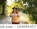 Stylish 30s woman holding paper map relaxing in nature. Travel and active lifestyle concept. Happy female lifestyle wearing backpack and hat exercising outdoors. 143695457