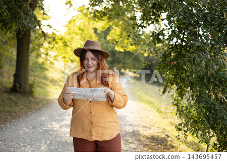 Stylish 30s woman holding paper map relaxing in nature. Travel and active lifestyle concept. Happy female lifestyle wearing backpack and hat exercising outdoors. 143695457