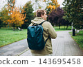 A man with backpack walking on the street in the city park. Urban lifestyle concept. Traveler. Autumn time. Young male in casual clothing standing outdoor. Rear view 143695458