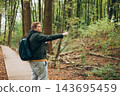 Blonde young 30s man wearing backpack holding phone and pointing something on city autumn park street. Urban lifestyle, travel concept. High quality photo 143695459