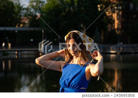 Portrait of woman on sunset in green par wearing deep blue dress and knitted little scarf on head illustrating summer warm atmosphere and freedom 143695808