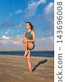 A woman prepares for Utthita Hasta Padangusthasana yoga, Extended Hand-to-Big-Toe Pose, balancing on one leg while hugging a bent knee on a beach 143696008