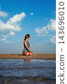 A woman practices yoga in a Bharadvajasana variation, a kneeling Bharadvaja Twist, turning her torso while seated on her heels on beach sand 143696010