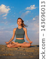 Facing forward on the beach, a woman holds Padmasana yoga, Lotus Pose, with closed eyes and upturned palms resting on her knees under a blue sky 143696013