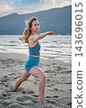 A woman holds Virabhadrasana 2 yoga, Warrior 2 Pose, bending her front knee and extending both arms level in a three-quarter beach view near a coastal hill 143696015