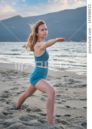 A woman holds Virabhadrasana 2 yoga, Warrior 2 Pose, bending her front knee and extending both arms level in a three-quarter beach view near a coastal hill A woman holds Virabhadrasana 2 yoga, Warrior 2 Pose, bending her front knee and extending both arms level in a three-quarter beach view near a coastal hill 143696015