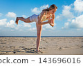 A woman balances in a Virabhadrasana 3 yoga variation, Warrior 3 Pose, leaning forward with prayer hands and a slightly bent raised back leg on beach sand 143696016