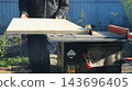 A worker dressed in dark clothing carefully feeds a wide wooden furniture panel through the rotating saw blade of a table saw. The board is being ripped lengthwise outdoors on a sunny day. 143696405