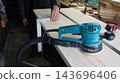 A craftsman measures a smooth wooden board using a metal carpenters square in a woodworking shop. In the foreground, an orbital sander lies on the surface, connected to a vacuum cleaner hose. 143696406