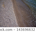 Top-down view of a gravelly pebble beach meeting clear water. Gentle waves lap against the dark, wet shoreline, creating a distinct natural line between dry small stones and wet sand. 143696632