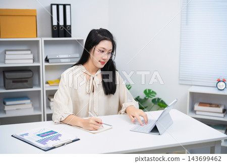 Young Asian Woman Working with Tablet and Notepad in Modern Office, Analyzing Data and Making Notes, Flanked by Shelves and Office Supplies Young Asian Woman Working with Tablet and Notepad in Modern Office, Analyzing Data and Making Notes, Flanked by Shelves and Office Supplies 143699462