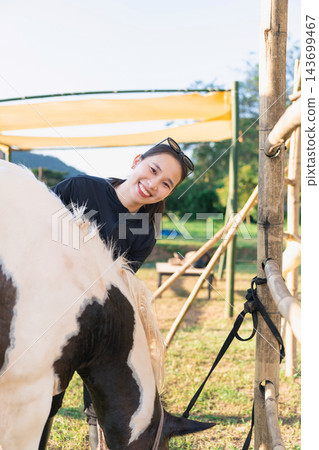 Young woman smiling and interacting with horse outdoors in sunny rural area with wooden fence and green nature background Young woman smiling and interacting with horse outdoors in sunny rural area with wooden fence and green nature background 143699467