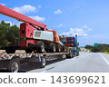 Blue truck carries red boom lift on flatbed trailer along highway in daylight. 143699621