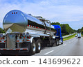 Blue cab pulls stainless steel tanker truck along highway beside trees, traffic. 143699622