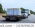 Pickup truck hauls flatbed trailer carrying containers along road near trees, buildings in daylight. 143699626