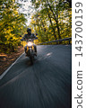 Motorcyclist wearing a black helmet and dark riding gear approaching the camera on a winding asphalt road through a sunlit green and golden autumn forest, with dynamic motion blur. 143700159