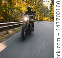 Motorcyclist wearing a black helmet and dark riding gear approaching the camera on a winding asphalt road through a sunlit green and golden autumn forest, with dynamic motion blur. 143700160