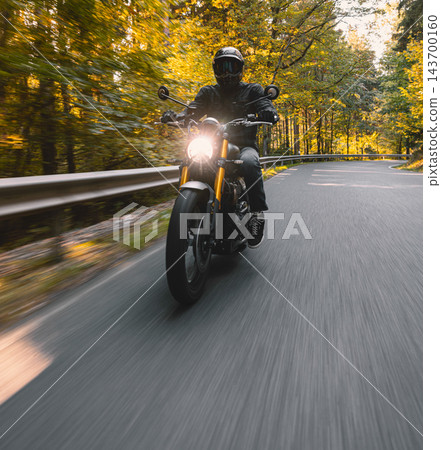 Motorcyclist wearing a black helmet and dark riding gear approaching the camera on a winding asphalt road through a sunlit green and golden autumn forest, with dynamic motion blur. 143700160