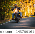 Low angle action photograph of a motorcyclist in black protective gear leaning into a curve on an asphalt road surrounded by golden autumn trees, with warm sunlight and motion blur. 143700161
