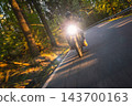 Motorcyclist wearing a black helmet and dark riding gear approaching the camera on a winding asphalt road through a sunlit green and golden autumn forest, with dynamic motion blur. 143700163