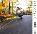 Motorcyclist wearing a black helmet and dark riding gear approaching the camera on a winding asphalt road through a sunlit green and golden autumn forest, with dynamic motion blur. 143700165