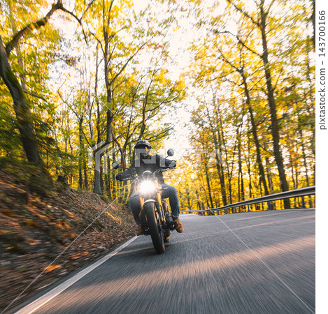 Motorcyclist wearing a black helmet and dark riding gear approaching the camera on a winding asphalt road through a sunlit green and golden autumn forest, with dynamic motion blur. Motorcyclist wearing a black helmet and dark riding gear approaching the camera on a winding asphalt road through a sunlit green and golden autumn forest, with dynamic motion blur. 143700166