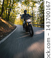 Motorcyclist wearing a black helmet and dark riding gear approaching the camera on a winding asphalt road through a sunlit green and golden autumn forest, with dynamic motion blur. 143700167
