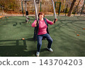 Mature woman exercising outdoors on public fitness equipment. Active retirement and physical wellbeing. 143704203