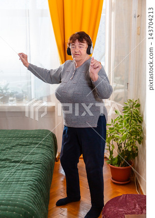 Older Caucasian woman listening to relaxing music and moving gently indoors. Calm mood and comfort. Concept of wellness for seniors. Vertical Older Caucasian woman listening to relaxing music and moving gently indoors. Calm mood and comfort. Concept of wellness for seniors. Vertical 143704213