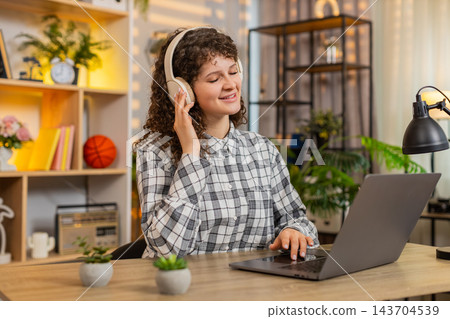 Young woman on home table typing on laptop in headphones listening to music during calm remote work Young woman on home table typing on laptop in headphones listening to music during calm remote work 143704539