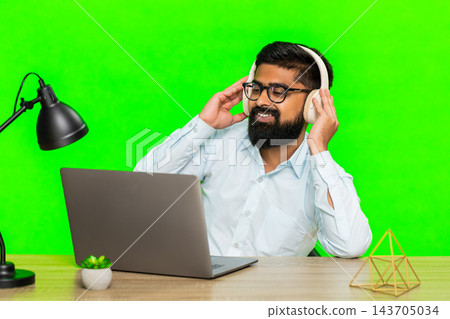 Indian man with wireless headphones working on laptop enjoying music nodding head listening focused Indian man with wireless headphones working on laptop enjoying music nodding head listening focused 143705034
