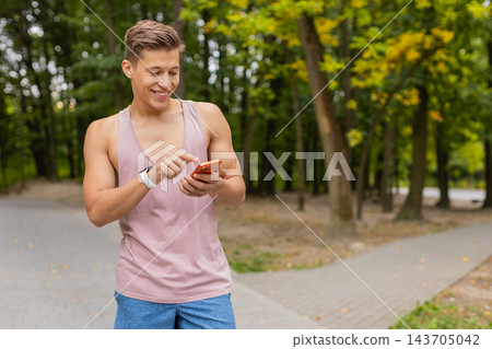 Young man smiling down at orange smartphone while wearing pink tank top and smartwatch in city park 143705042