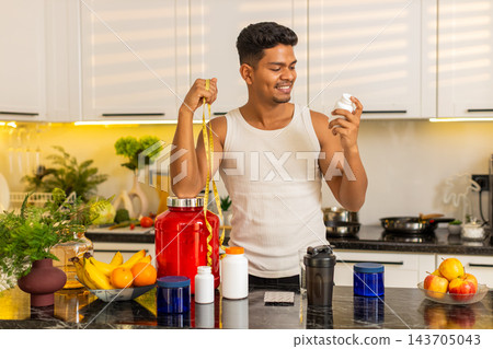 Indian man approving sports supplement with measuring tape for fitness diet progress in home kitchen 143705043