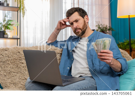 Man holding cash dollars in hands thoughtfully looking at laptop screen deciding how to spend money Man holding cash dollars in hands thoughtfully looking at laptop screen deciding how to spend money 143705061