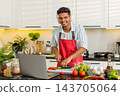 Indian man slicing cucumber for fresh salad while watching laptop in kitchen for healthy meal prep 143705064