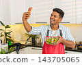 Indian man showing vegetable salad to smartphone while sharing healthy meal tips in modern kitchen 143705065