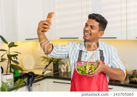 Indian man showing vegetable salad to smartphone while sharing healthy meal tips in modern kitchen Indian man showing vegetable salad to smartphone while sharing healthy meal tips in modern kitchen 143705065