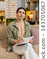 Young woman on home sofa writing plans in notebook with earbuds while studying and thinking indoors 143705067