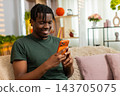African American young man on home sofa reading smartphone message smiling in cozy living room chat 143705075