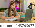 Child girl kid doing online homework on laptop and writing math notes while sitting at home table 143705077