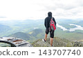 Hiker with red backpack standing on rocky summit above expansive green mountain valley and winding lakes in a scenic wilderness landscape 143707740